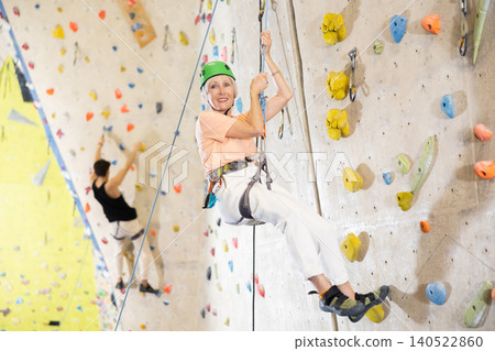 Mature woman in safety gear doing difficult wall climb in climbing gym Mature woman in safety gear doing difficult wall climb in climbing gym 140522860
