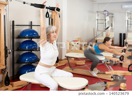 Sports club senior woman client performs exercise on reformer with cable and rope for arms Sports club senior woman client performs exercise on reformer with cable and rope for arms 140523679