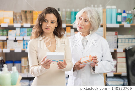 Woman choose antiallergic pills in pharmacy, mature woman pharmacist in background Woman choose antiallergic pills in pharmacy, mature woman pharmacist in background 140523709