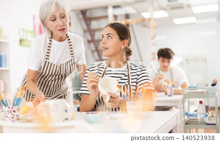 Senior woman assisting young girl painting ceramic mug in pottery class Senior woman assisting young girl painting ceramic mug in pottery class 140523938