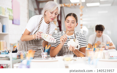 Senior woman assisting young girl painting ceramic mug in pottery class 140523957