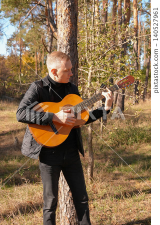 Portrait of smiling caucasian artist plays wooden acoustic guitar in autumnal park. Young songwriter plays string musical instrument hobby outside in nature fall time. Audio music healing Portrait of smiling caucasian artist plays wooden acoustic guitar in autumnal park. Young songwriter plays string musical instrument hobby outside in nature fall time. Audio music healing 140527961