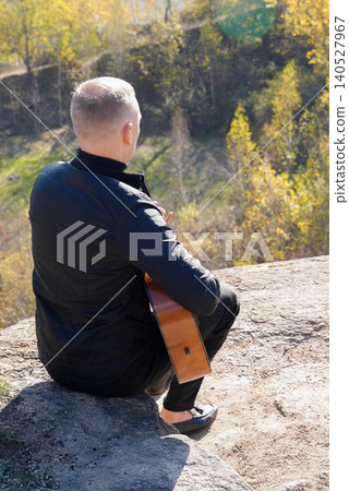 Beautiful blonde man playing acoustic guitar and singing relaxing enjoying outdoors on sunny autumn day. Artist learning and practicing playing musical instrument. Fall leaves autumnal 140527967