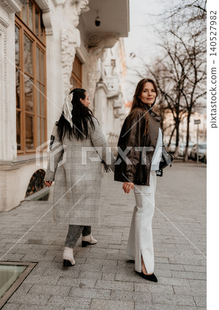 Women fashion city stroll as two stylish young adults walk along an urban sidewalk wearing autumn jackets and coats Women fashion city stroll as two stylish young adults walk along an urban sidewalk wearing autumn jackets and coats 140527982