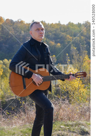 Blonde man playing string guitar outdoors in autumn forest. Concept of sound therapy, mental health and wellness rituals. Calmness tranquility audio-sensory practices. Aura farming energy Blonde man playing string guitar outdoors in autumn forest. Concept of sound therapy, mental health and wellness rituals. Calmness tranquility audio-sensory practices. Aura farming energy 140528000