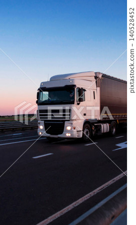 White semi-truck with dark cargo trailer travels highway at twilight, sky gradient blue to pink horizon, headlights illuminated, long haul transportation vehicle movement, vertical photo 140528452