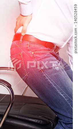 Person experiencing lower back pain or hip discomfort, hand holding painful region of body while sitting on office chair edge, wearing white shirt, blue jeans, red belt, red highlight indicates Person experiencing lower back pain or hip discomfort, hand holding painful region of body while sitting on office chair edge, wearing white shirt, blue jeans, red belt, red highlight indicates 140528459