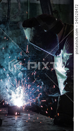 Skilled worker performing arc welding process with intense bright sparks and swirling blue smoke in dark industrial workshop, wearing full protective helmet and gear, close-up action vertical photo Skilled worker performing arc welding process with intense bright sparks and swirling blue smoke in dark industrial workshop, wearing full protective helmet and gear, close-up action vertical photo 140528472