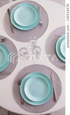 Elegant dining table set for three with aquamarine plates, grey woven placemats, and polished silverware, clear empty glasses on white surface, overhead view vertical photo Elegant dining table set for three with aquamarine plates, grey woven placemats, and polished silverware, clear empty glasses on white surface, overhead view vertical photo 140528476