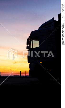 Silhouette of large semi-truck against vibrant sunset sky with shades of orange, yellow, and purple, featuring distant horizon and fence, vertical photo 140528494
