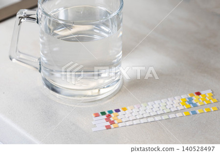 Water quality analysis using multiple test strips next to clear glass mug filled with pure water on clean light-colored surface, illustrating detailed chemical testing of liquid purity Water quality analysis using multiple test strips next to clear glass mug filled with pure water on clean light-colored surface, illustrating detailed chemical testing of liquid purity 140528497