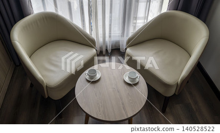 Top view of two cream leather armchairs with two white coffee cups on a round wooden table. Sheer curtains and dark floor in a cozy hotel room in Vietnam. Top view of two cream leather armchairs with two white coffee cups on a round wooden table. Sheer curtains and dark floor in a cozy hotel room in Vietnam. 140528522