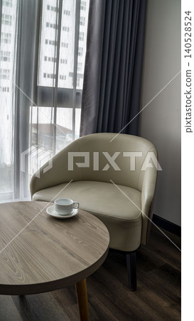Elegant cream leather armchair next to a round wooden table with a white coffee cup. Sheer curtains, soft natural light, and city view in a modern hotel room in Vietnam. Elegant cream leather armchair next to a round wooden table with a white coffee cup. Sheer curtains, soft natural light, and city view in a modern hotel room in Vietnam. 140528524