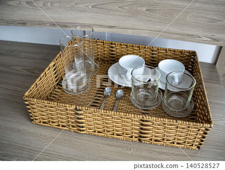 Wicker tray with glass cups, white ceramic coffee cups with saucers, and teaspoons on a wooden surface in a hotel room. Neat hospitality amenities set. 140528527