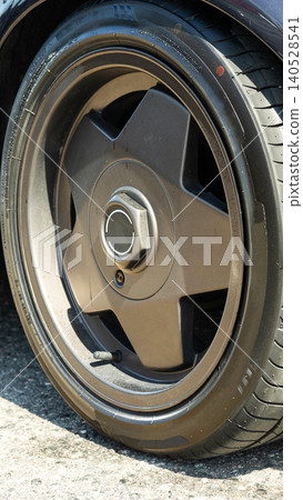 Close-up of car wheel and tire on asphalt during day, focus on customized dark multi-spoke alloy rim, detailed view of tire tread and sidewall, vertical shot of vehicle component Close-up of car wheel and tire on asphalt during day, focus on customized dark multi-spoke alloy rim, detailed view of tire tread and sidewall, vertical shot of vehicle component 140528541