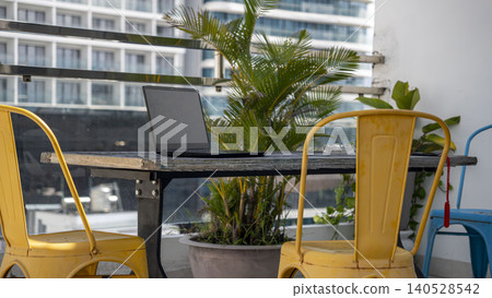 Laptop on an industrial table on a rooftop terrace in Vietnam. Yellow metal chair, tropical plants, glass railing, and city skyline with mountains. Digital nomad lifestyle. Laptop on an industrial table on a rooftop terrace in Vietnam. Yellow metal chair, tropical plants, glass railing, and city skyline with mountains. Digital nomad lifestyle. 140528542
