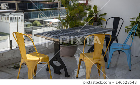 Stylish rooftop terrace in Vietnam with colorful metal chairs in yellow, black, and blue around an industrial table. Tropical plants, glass railing, and city skyline with mountains. 140528548