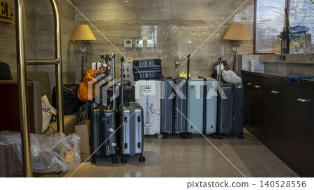 Multiple suitcases and travel bags lined up in a hotel marble lobby storage area. Various sizes and colors of luggage, backpacks, table lamps, and a luggage cart visible on the side. Multiple suitcases and travel bags lined up in a hotel marble lobby storage area. Various sizes and colors of luggage, backpacks, table lamps, and a luggage cart visible on the side. 140528556
