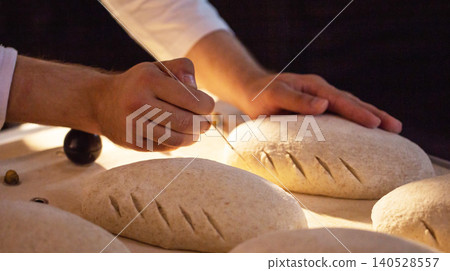 Hands scoring raw bread dough loaves with sharp tool before baking process in artisan bakery kitchen preparing traditional handmade bread products 140528557