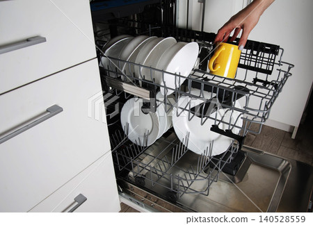 Human hand loading dirty dishes into modern automatic dishwasher kitchen appliance. White ceramic plates yellow mug organized inside kitchen dishwasher. Human hand loading dirty dishes into modern automatic dishwasher kitchen appliance. White ceramic plates yellow mug organized inside kitchen dishwasher. 140528559