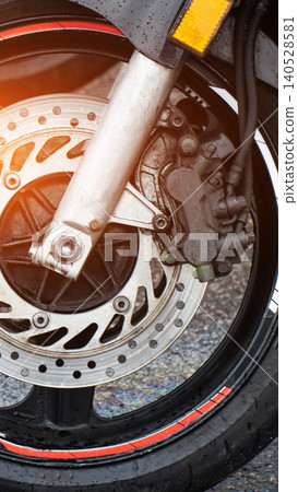 Close-up detail of motorcycle front wheel with disc brake system and suspension fork. Wet conditions visible on fender and tire. Sunlight lens flare illuminates mechanical components. 140528581