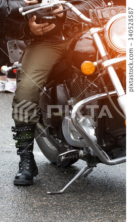 Person sitting motorcycle holding phone wearing black leather jacket green pants black boots with spikes buckles on wet asphalt foreground motorcycle headlight engine chrome detail vertical photo Person sitting motorcycle holding phone wearing black leather jacket green pants black boots with spikes buckles on wet asphalt foreground motorcycle headlight engine chrome detail vertical photo 140528587