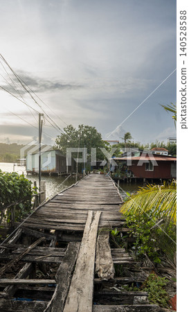 A crumbling wooden boardwalk leads through a remote Vietnamese fishing village with stilt houses, palm trees and power lines under a pale cloudy sky. Vietnam. A crumbling wooden boardwalk leads through a remote Vietnamese fishing village with stilt houses, palm trees and power lines under a pale cloudy sky. Vietnam. 140528588