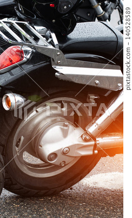 Black motorcycle rear wheel detail, wet from rain, with chrome exhaust pipes, fender, taillight, and turn signal, parked on asphalt, vertical shot. Black motorcycle rear wheel detail, wet from rain, with chrome exhaust pipes, fender, taillight, and turn signal, parked on asphalt, vertical shot. 140528589