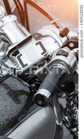 Close-up view of wet motorcycle handlebars and fuel tank covered with raindrops, chrome details reflecting light, smartphone holder mounted, classic cruiser parked outdoors Close-up view of wet motorcycle handlebars and fuel tank covered with raindrops, chrome details reflecting light, smartphone holder mounted, classic cruiser parked outdoors 140528592