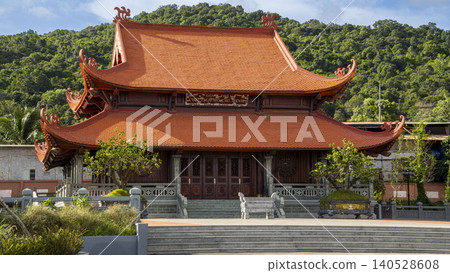 Side-angle view of the Ho Chi Minh memorial temple in Vietnam showing intricate wooden facade and sweeping traditional orange tiled roof against forested hills. Side-angle view of the Ho Chi Minh memorial temple in Vietnam showing intricate wooden facade and sweeping traditional orange tiled roof against forested hills. 140528608