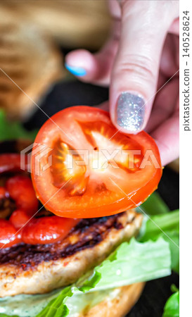 Woman hand placing fresh ripe tomato slice on grilled chicken sandwich with vibrant green lettuce and creamy sauce, preparing delicious meal close up vertical photo 140528624