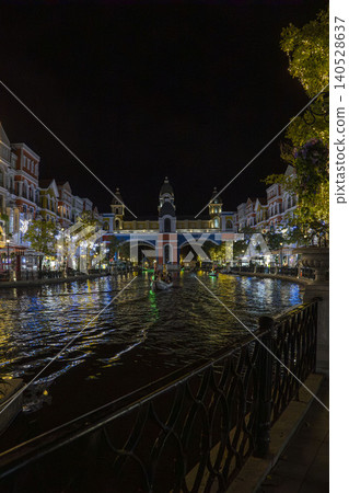 Night view down a glowing Venice-style canal toward an ornate illuminated arch bridge with gondola, colorful European buildings and festive lights in Phu Quoc, Vietnam. Night view down a glowing Venice-style canal toward an ornate illuminated arch bridge with gondola, colorful European buildings and festive lights in Phu Quoc, Vietnam. 140528637