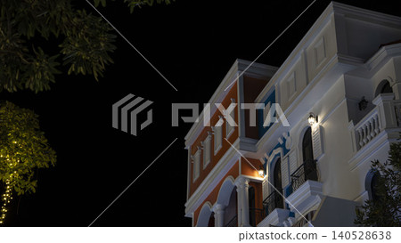 Low-angle night view of an elegant European-style building facade with orange walls, white arches and balconies lit by wall lanterns against a dark sky in Phu Quoc, Vietnam. Low-angle night view of an elegant European-style building facade with orange walls, white arches and balconies lit by wall lanterns against a dark sky in Phu Quoc, Vietnam. 140528638