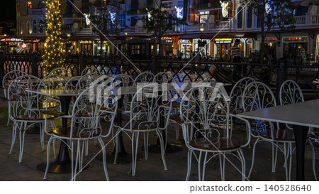 Empty white wrought iron cafe chairs and tables beside a festively lit canal with European-style buildings at night in Grand World, Phu Quoc, Vietnam. Empty white wrought iron cafe chairs and tables beside a festively lit canal with European-style buildings at night in Grand World, Phu Quoc, Vietnam. 140528640