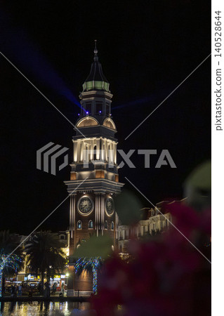 Majestic illuminated clock tower with blue spotlights rising into the dark night sky at Grand World entertainment complex in Phu Quoc, Vietnam. Bokeh foreground. Majestic illuminated clock tower with blue spotlights rising into the dark night sky at Grand World entertainment complex in Phu Quoc, Vietnam. Bokeh foreground. 140528644
