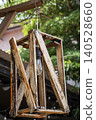 A handcrafted wooden lantern with a geometric frame of weathered planks hangs from a rope in an outdoor garden setting. A vintage-style filament bulb glows softly at its centre. 140528660