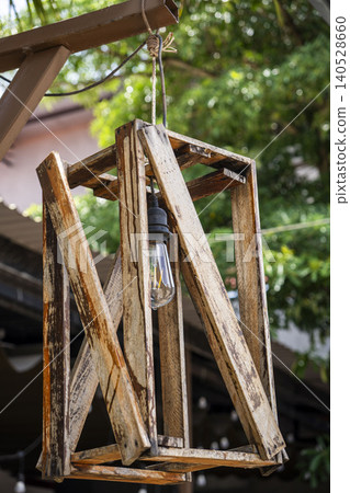A handcrafted wooden lantern with a geometric frame of weathered planks hangs from a rope in an outdoor garden setting. A vintage-style filament bulb glows softly at its centre. A handcrafted wooden lantern with a geometric frame of weathered planks hangs from a rope in an outdoor garden setting. A vintage-style filament bulb glows softly at its centre. 140528660