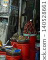 A Vietnamese street food stall with red plastic buckets used as improvised counters, displaying piles of dried sausages, pickled vegetables, and preserved foods in metal bowls. Vietnamese signage 140528661