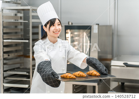 一位身穿廚師服的女廚師在廚房裡手捧一個新鮮出爐的羊角麵包。 一位身穿廚師服的女廚師在廚房裡手捧一個新鮮出爐的羊角麵包。 140528712