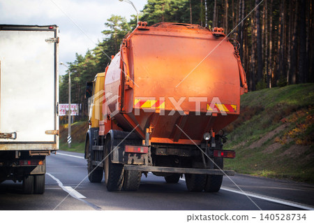 Rear view of orange garbage truck and white truck driving on asphalt road beside forest slope on overcast day 140528734