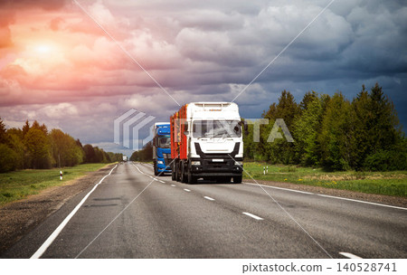 Heavy white truck transports logs on rural highway, blue semi truck follows, green forest lines road, cloudy sky above, long distance freight journey Heavy white truck transports logs on rural highway, blue semi truck follows, green forest lines road, cloudy sky above, long distance freight journey 140528741