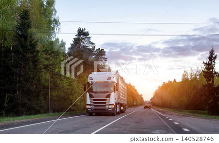 Large white semi truck driving on rural asphalt road through dense forest during sunset glow at evening time conveying heavy cargo 140528746