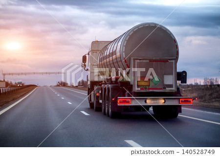 White tanker truck driving on asphalt highway at twilight, transporting hazardous liquid, rear view of vehicle on road under cloudy sky, evening sunset. Copy space for text 140528749