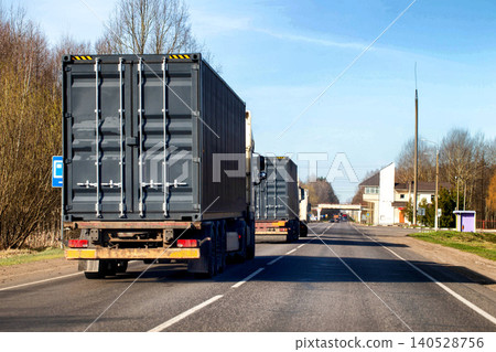 Two large semi-trailer trucks transport dark grey shipping containers on highway through rural landscape during day. Heavy cargo vehicles move on asphalt road. 140528756