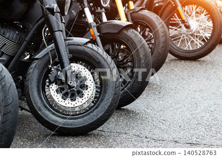 Row of parked motorcycles on wet asphalt road after rain, close view of front wheels, brake discs, and fenders, gleaming metal parts under bright outdoor light Row of parked motorcycles on wet asphalt road after rain, close view of front wheels, brake discs, and fenders, gleaming metal parts under bright outdoor light 140528763