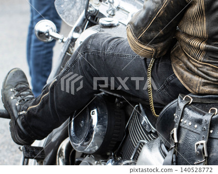 Person in black jeans, worn brown leather jacket, sturdy black boots sits on powerful cruiser motorcycle, close view of engine, chrome details, black leather saddlebag with buckles, golden chain Person in black jeans, worn brown leather jacket, sturdy black boots sits on powerful cruiser motorcycle, close view of engine, chrome details, black leather saddlebag with buckles, golden chain 140528772