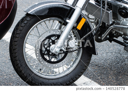 Front wheel of motorcycle with disc brake and chrome spokes standing on wet asphalt road after rain, showing details of suspension, fender, and engine components. Front wheel of motorcycle with disc brake and chrome spokes standing on wet asphalt road after rain, showing details of suspension, fender, and engine components. 140528778