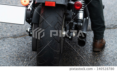 Rear view of motorcycle parked on wet asphalt road with person leg and brown shoe standing next to dual exhaust pipes and tail lights, license plate frame visible 140528780