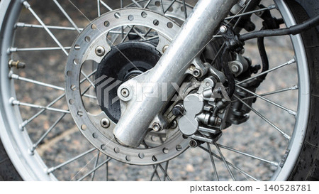 Close up view of front wheel assembly of modern motorcycle featuring disc brake system. Intricate detail of metal spokes, silver colored rim, functional suspension fork, wet surface. 140528781