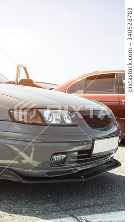 Front view of grey tuned car parked outdoors on bright sunny day, custom modifications visible on bumper and grille, second car partially visible in background, vertical photograph composition Front view of grey tuned car parked outdoors on bright sunny day, custom modifications visible on bumper and grille, second car partially visible in background, vertical photograph composition 140528783